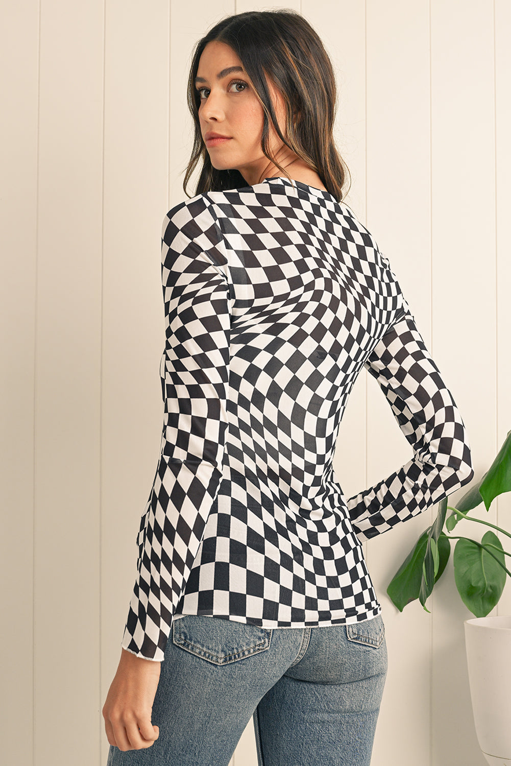 Woman wearing a black and white checkered long-sleeve top with a plant in the background.