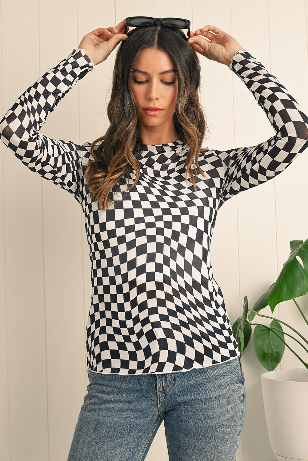 Woman wearing a black and white checkered long-sleeve top with a plant in the background.