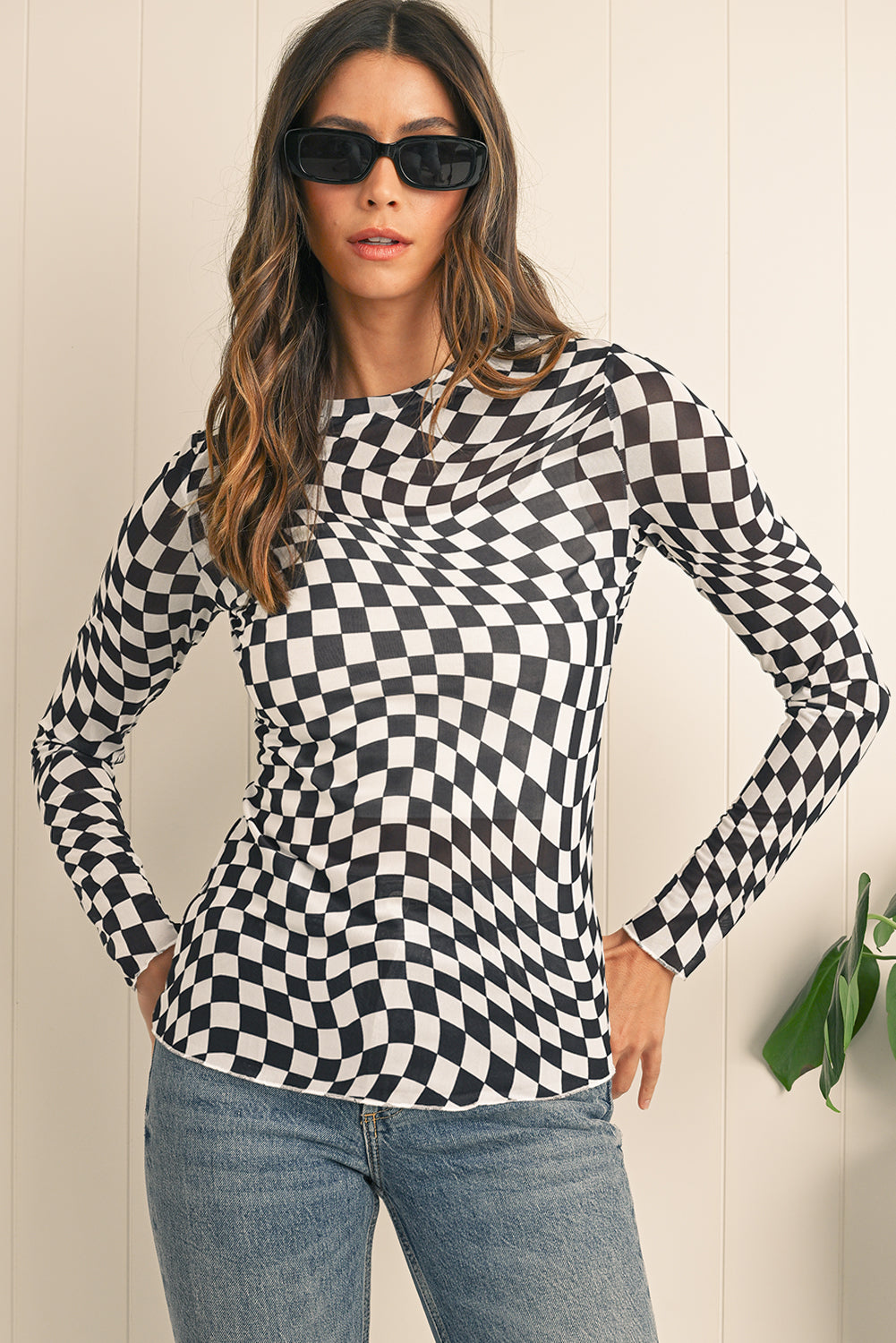Woman wearing a black and white checkered long-sleeve top with sunglasses against a light wooden panel background.