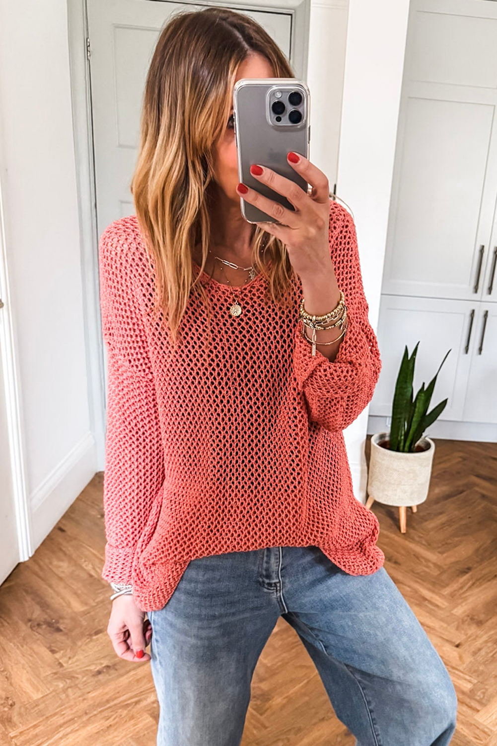 Woman taking a mirror selfie wearing a pink sweater and blue jeans in a room with wooden flooring and white cabinets.