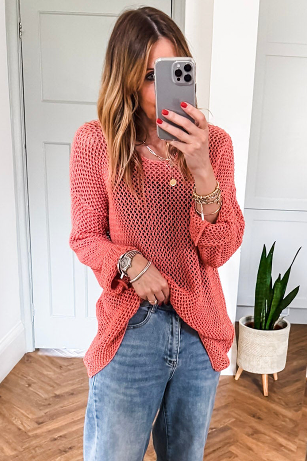 Woman taking a mirror selfie wearing a coral knitted sweater and blue jeans.
