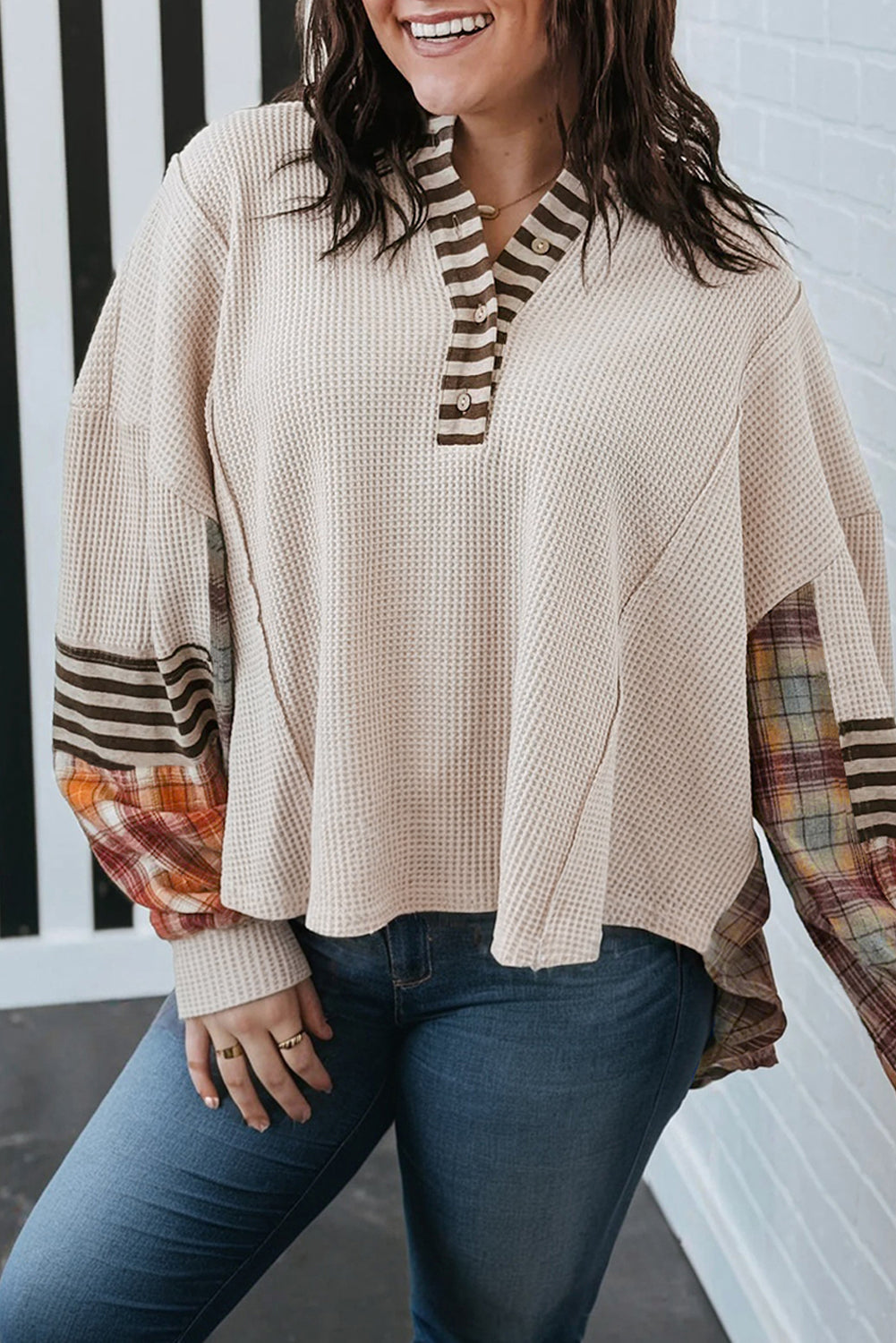 Woman wearing a beige poncho with plaid sleeves against a white wall.