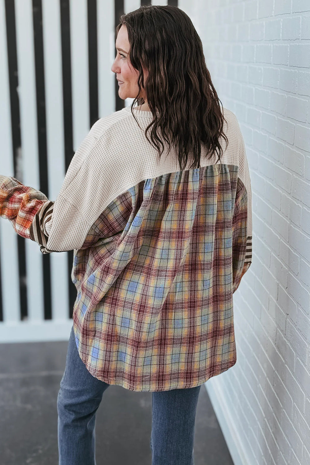 Woman wearing a plaid shirt over a beige sweater against a white brick wall.