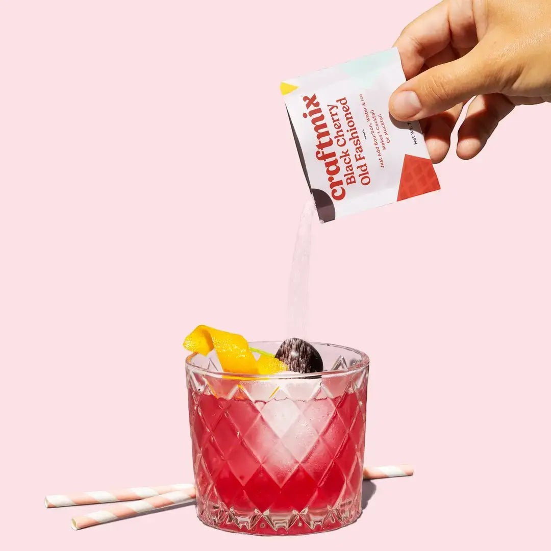 Doodlations Coffee Bar & Boutique Black Cherry Old Fashioned cocktail mix packet being poured into a diamond-cut glass with a cherry and orange peel garnish.