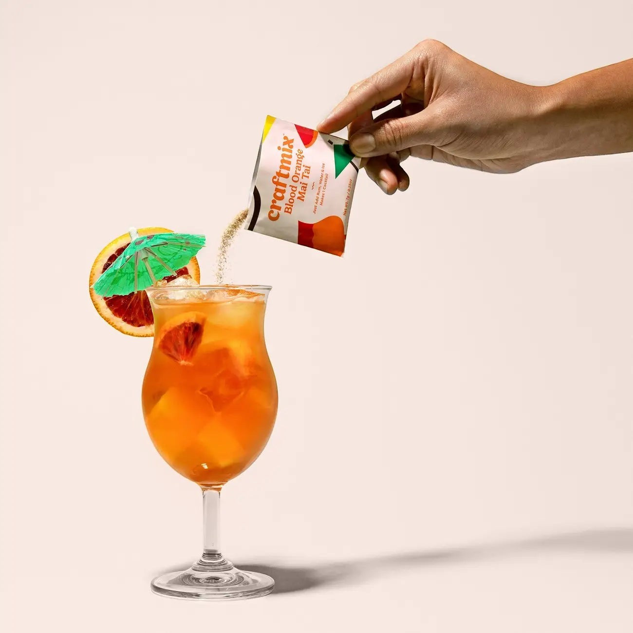 Doodlations Coffee Bar & Boutique Craftmix Blood Orange Mai Tai cocktail mix packet being poured into a hurricane glass with a tropical drink, ice, blood orange slices, and a cocktail umbrella.