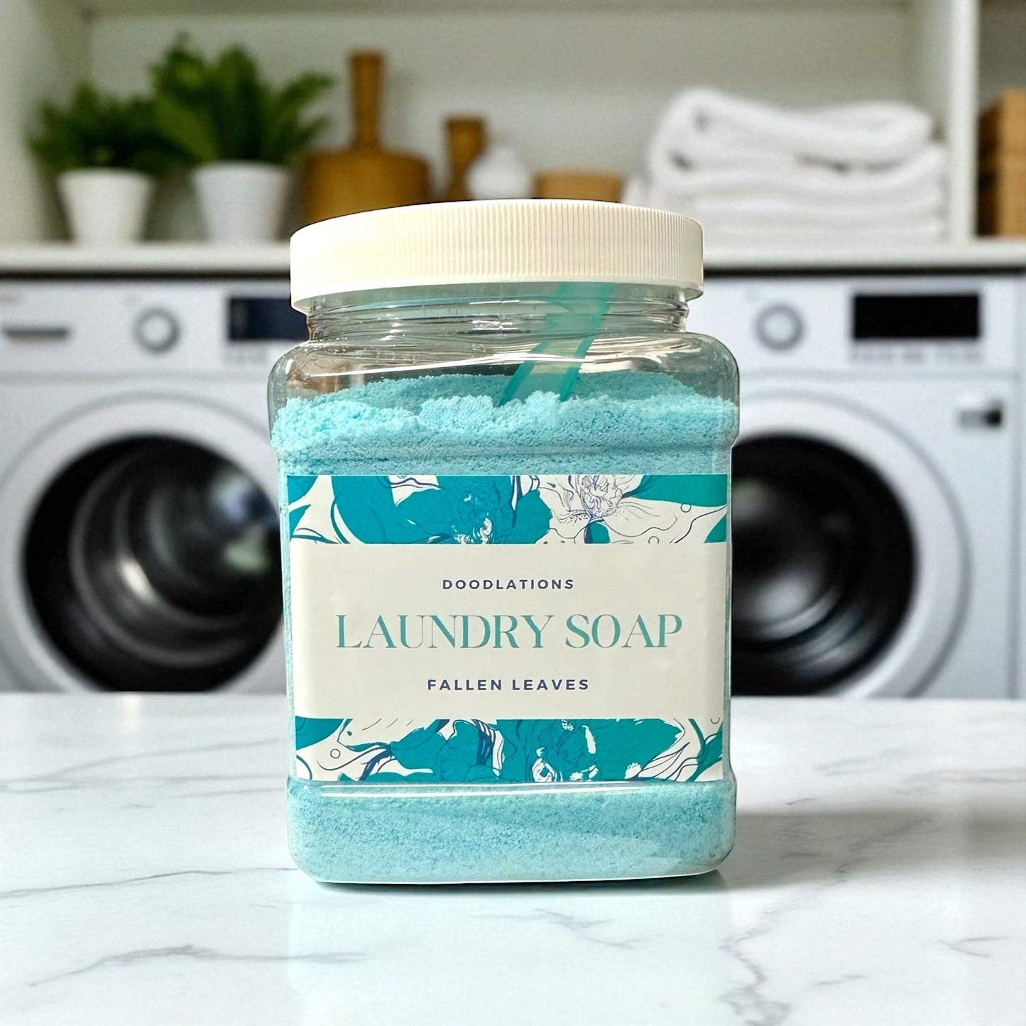 Doodlations Coffee Bar & Boutique Fallen Leaves powdered laundry detergent in a clear plastic jar with a blue scoop.