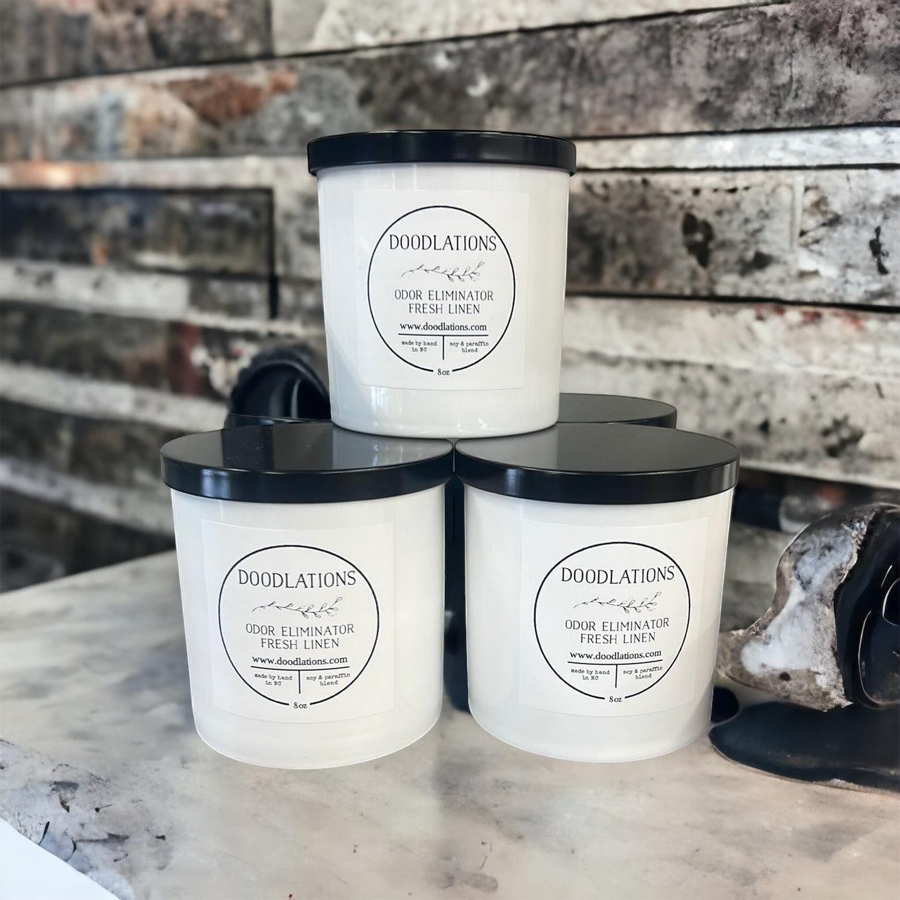 Doodlations Coffee Bar & Boutique Odor Eliminator luxury candle in fresh linen scent, white jar with black lid.