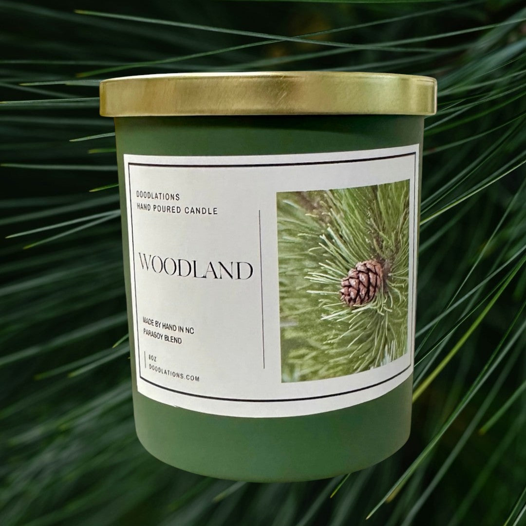 Doodlations Coffee Bar & Boutique Woodland hand-poured para-soy blend candle with pine cone imagery. Green jar, gold lid.