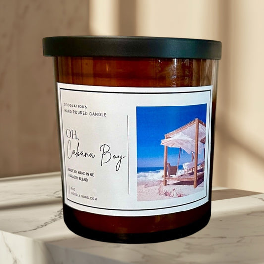 Doodlations Oh Cabana Boy hand-poured soy blend candle in amber glass with a black lid. Tropical beach scene on label.