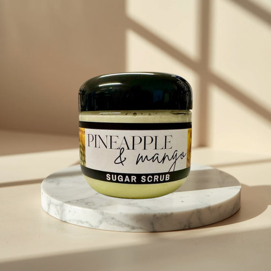 Pineapple Mango Sugar Scrub, a pale yellow exfoliating scrub in a clear jar with a black lid.