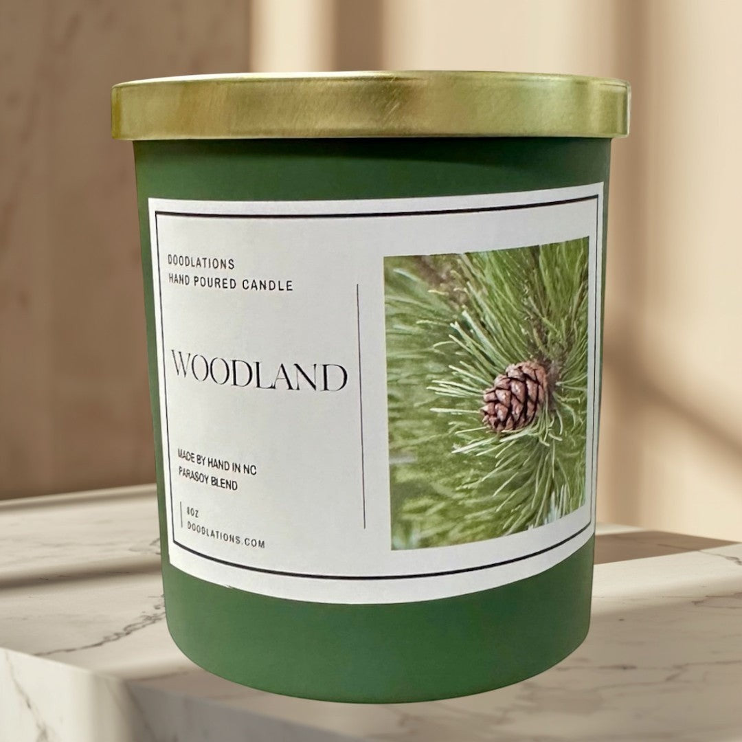 Woodland candle in a green jar with a gold lid, featuring a pine cone image.