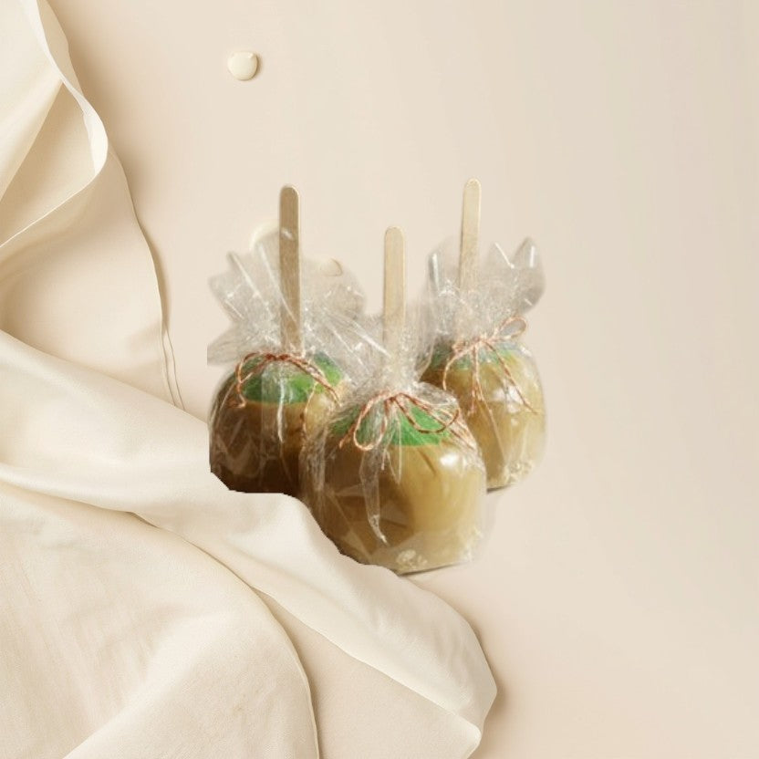 Three caramel apple wax melts with wooden sticks, wrapped in clear plastic and tied with red and green ribbons, displayed on a table.