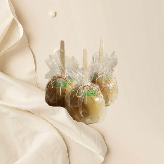 Three caramel apple wax melts with wooden sticks, wrapped in clear plastic and tied with red and green ribbons, displayed on a table.