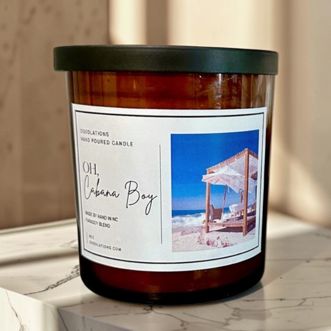 Doodlations Oh Cabana Boy hand-poured soy blend candle in amber glass with a black lid. Tropical beach scene on label.