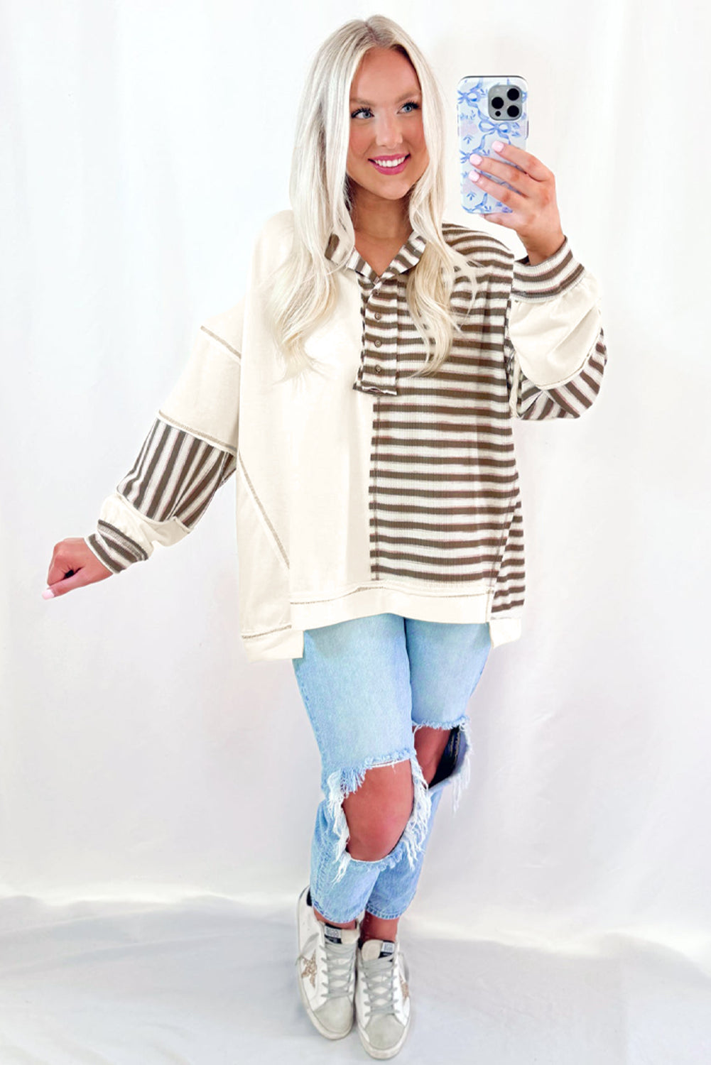 Woman wearing a striped sweater and ripped jeans, taking a selfie on a white background