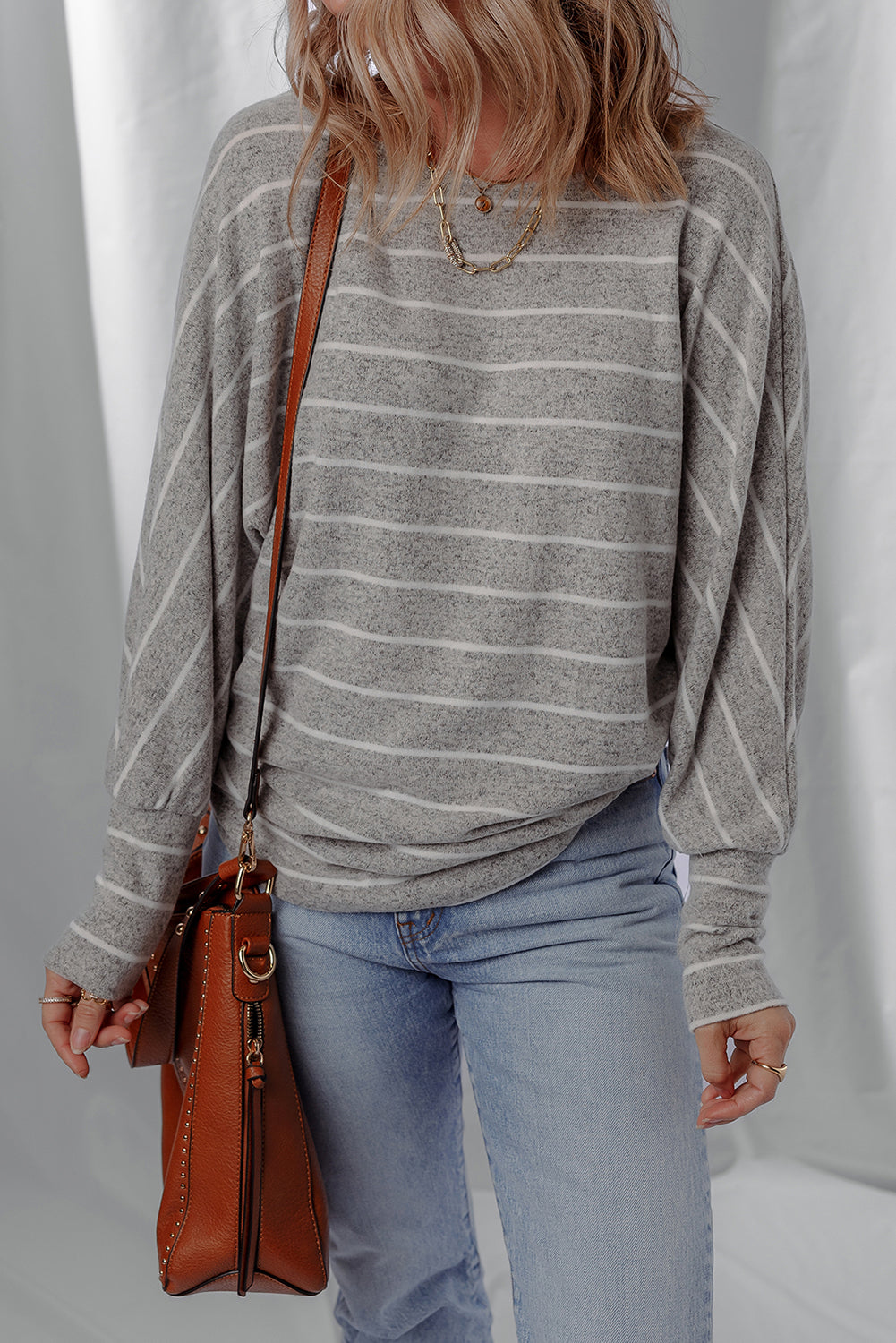 Person wearing a gray striped sweater and blue jeans holding a brown leather handbag against a white curtain background.