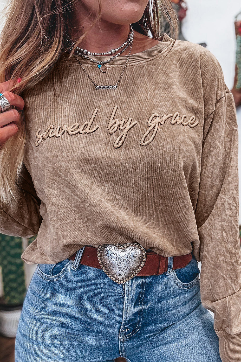Person wearing a brown sweatshirt with 'saved by grace' text, blue jeans, and a heart-shaped belt buckle.
