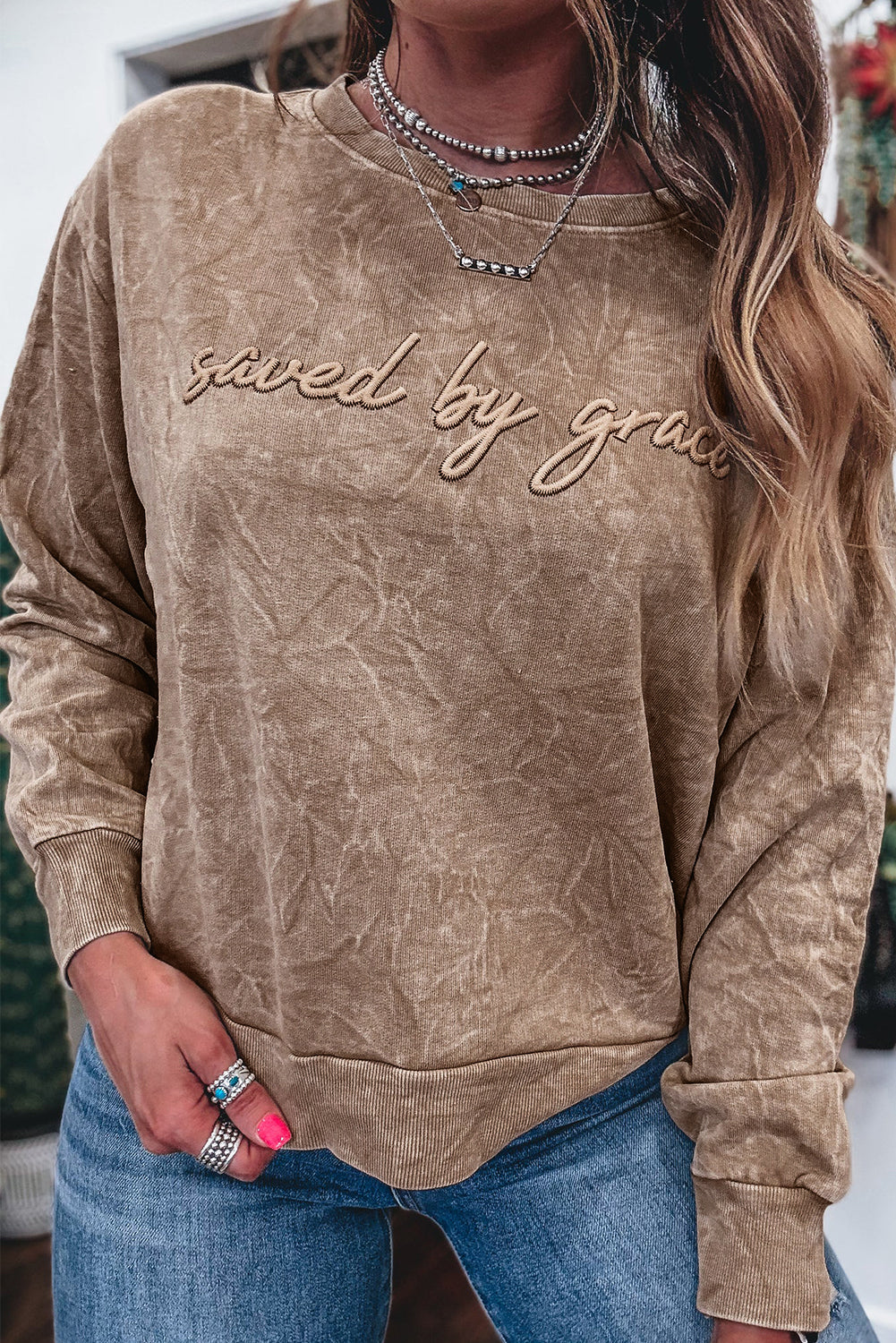 Person wearing a brown sweatshirt with 'saved by grace' text, holding jeans.