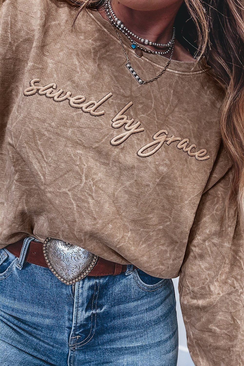 Brown distressed sweatshirt with 'saved by grace' text, worn with blue jeans and a belt.