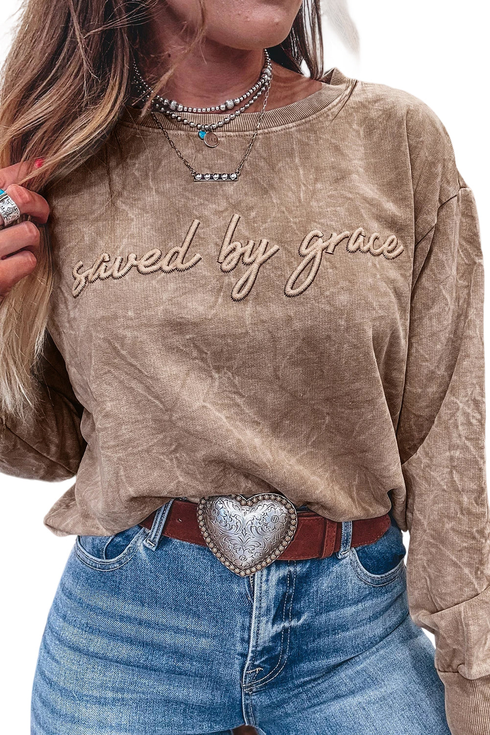 Person wearing a brown sweatshirt with 'saved by grace' text, blue jeans, and a heart-shaped belt buckle.