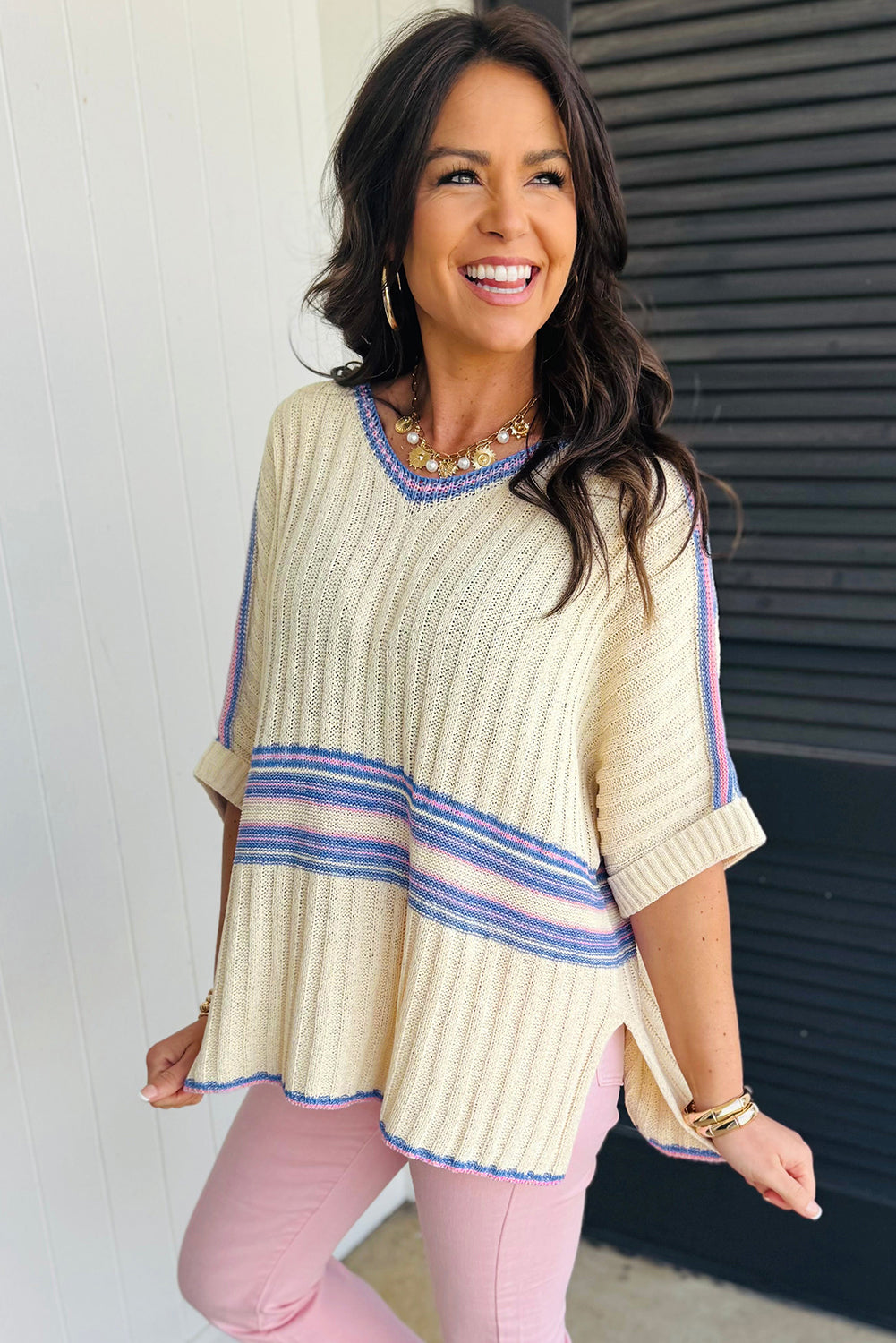 Woman wearing a light-colored sweater with blue stripes and pink pants against a white wall.