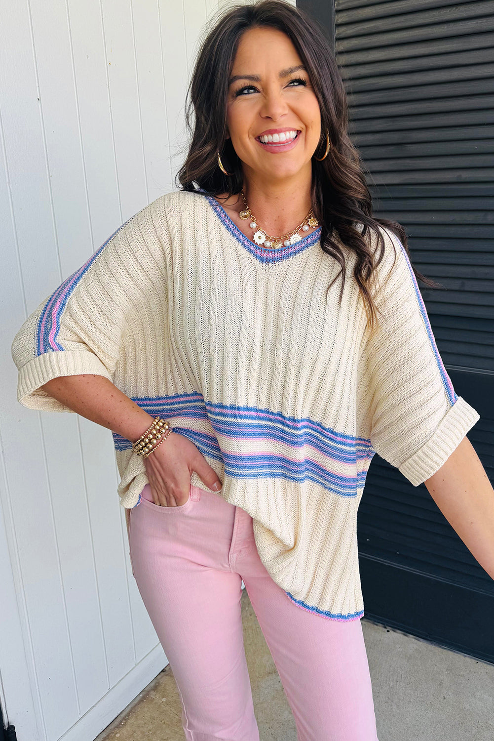 Woman wearing a cream sweater with blue stripes and pink pants standing against a white wall.