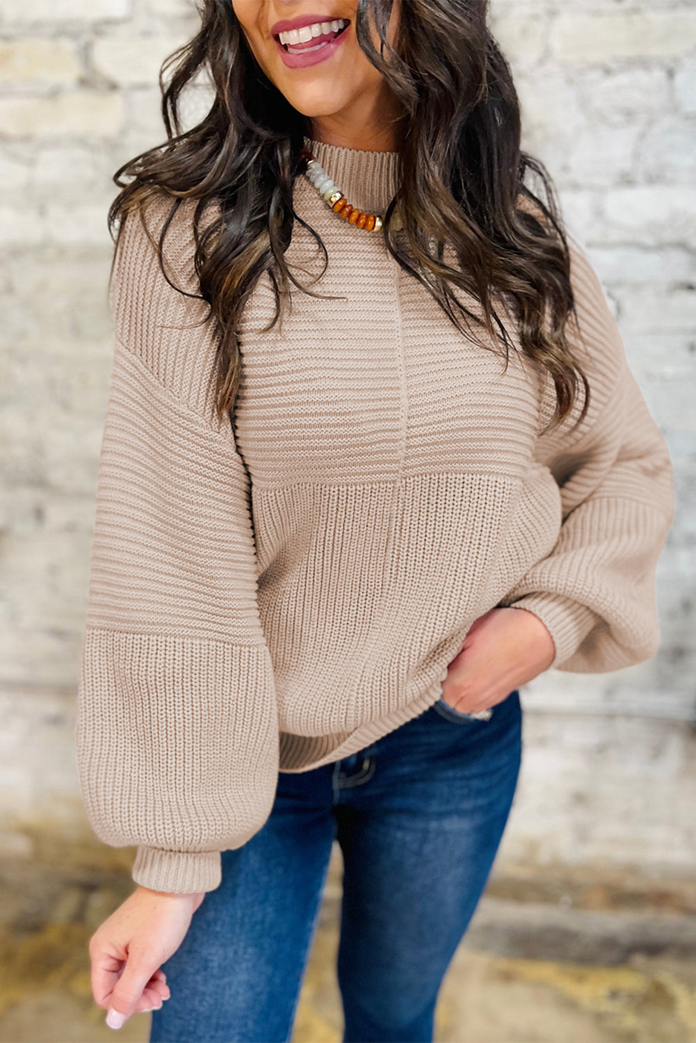 Woman wearing a beige sweater and blue jeans against a stone wall.