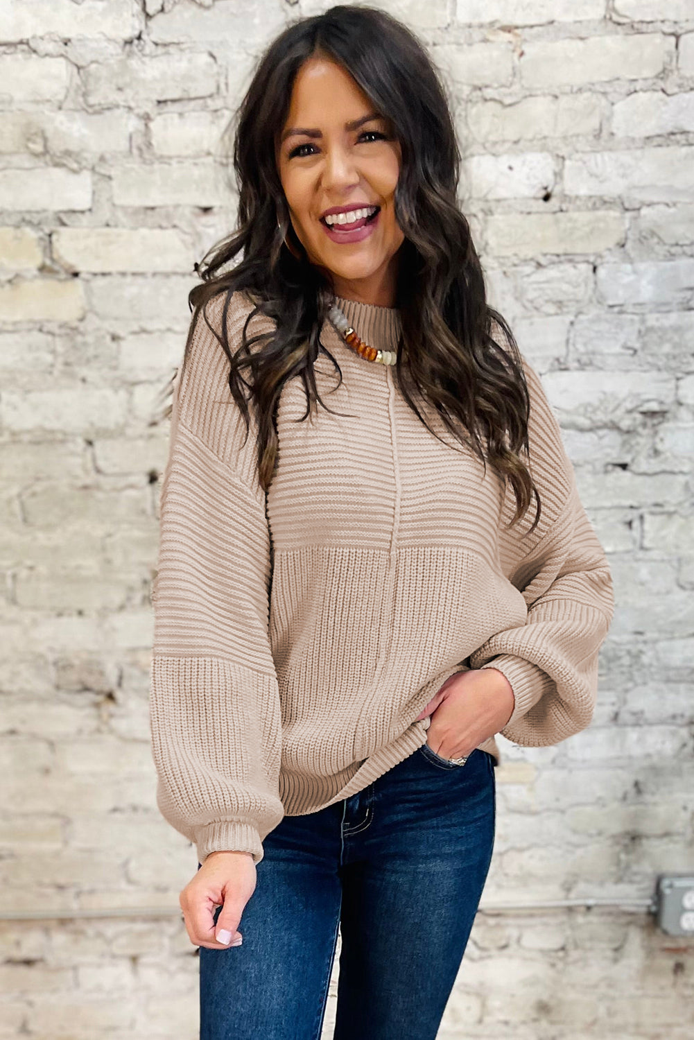 Woman wearing a beige sweater and blue jeans against a stone wall.