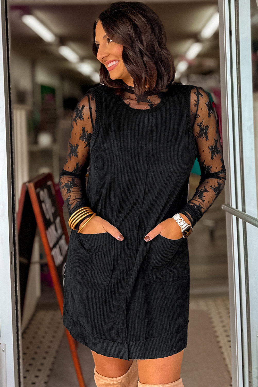 Woman wearing a black sleeveless dress with floral lace sleeves in a store setting.
