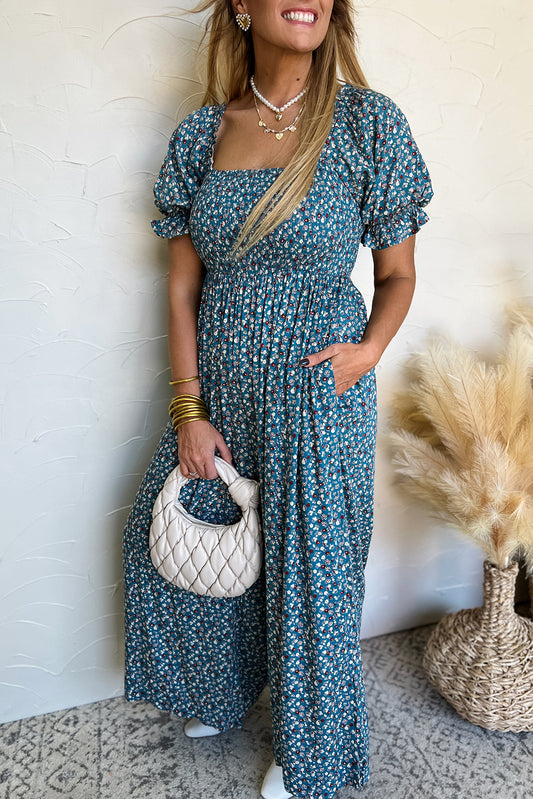 Woman in a blue floral dress holding a white handbag against a neutral background