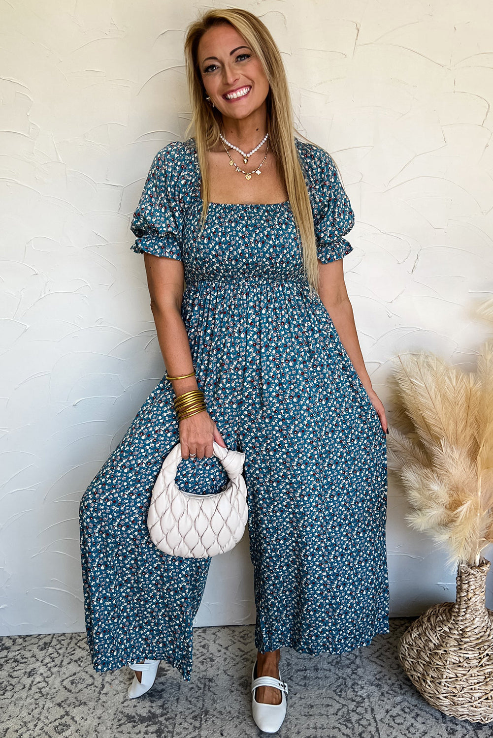 Woman wearing a blue floral dress holding a white handbag against a white wall.