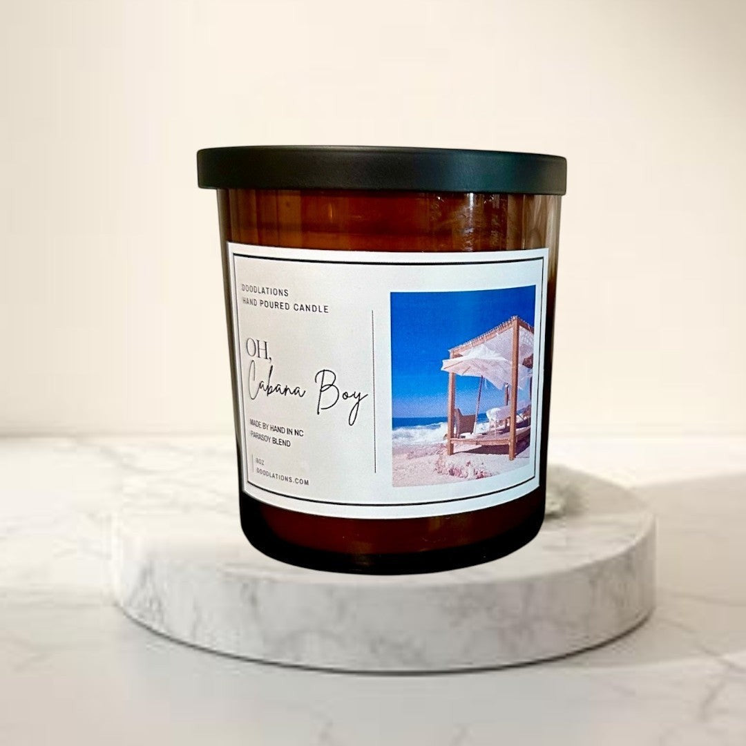 Doodlations Oh Cabana Boy hand-poured candle in amber glass with a black lid. Tropical beach scene on label.