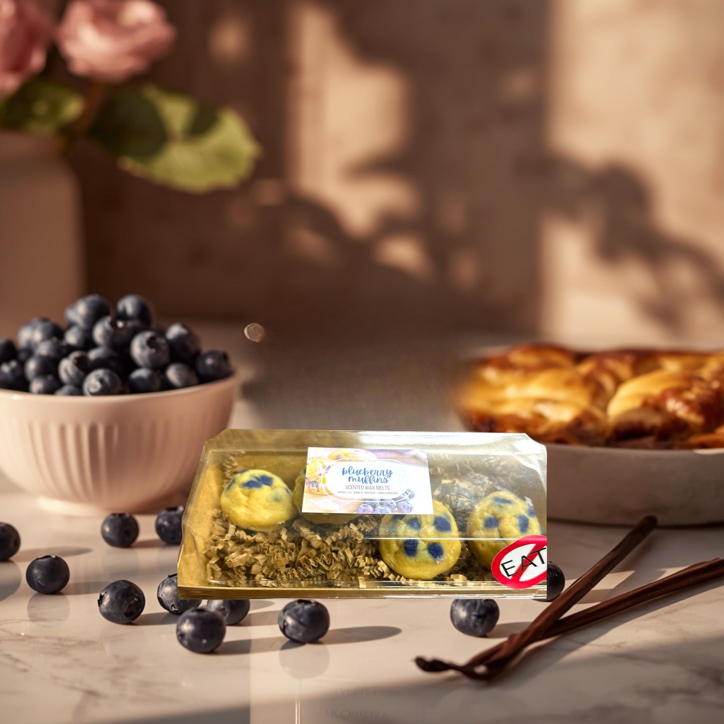 Doodlations Coffee Bar & Boutique Blueberry Muffin wax melts in a clear plastic container with shredded paper, on a striped cloth with greenery and white flowers.