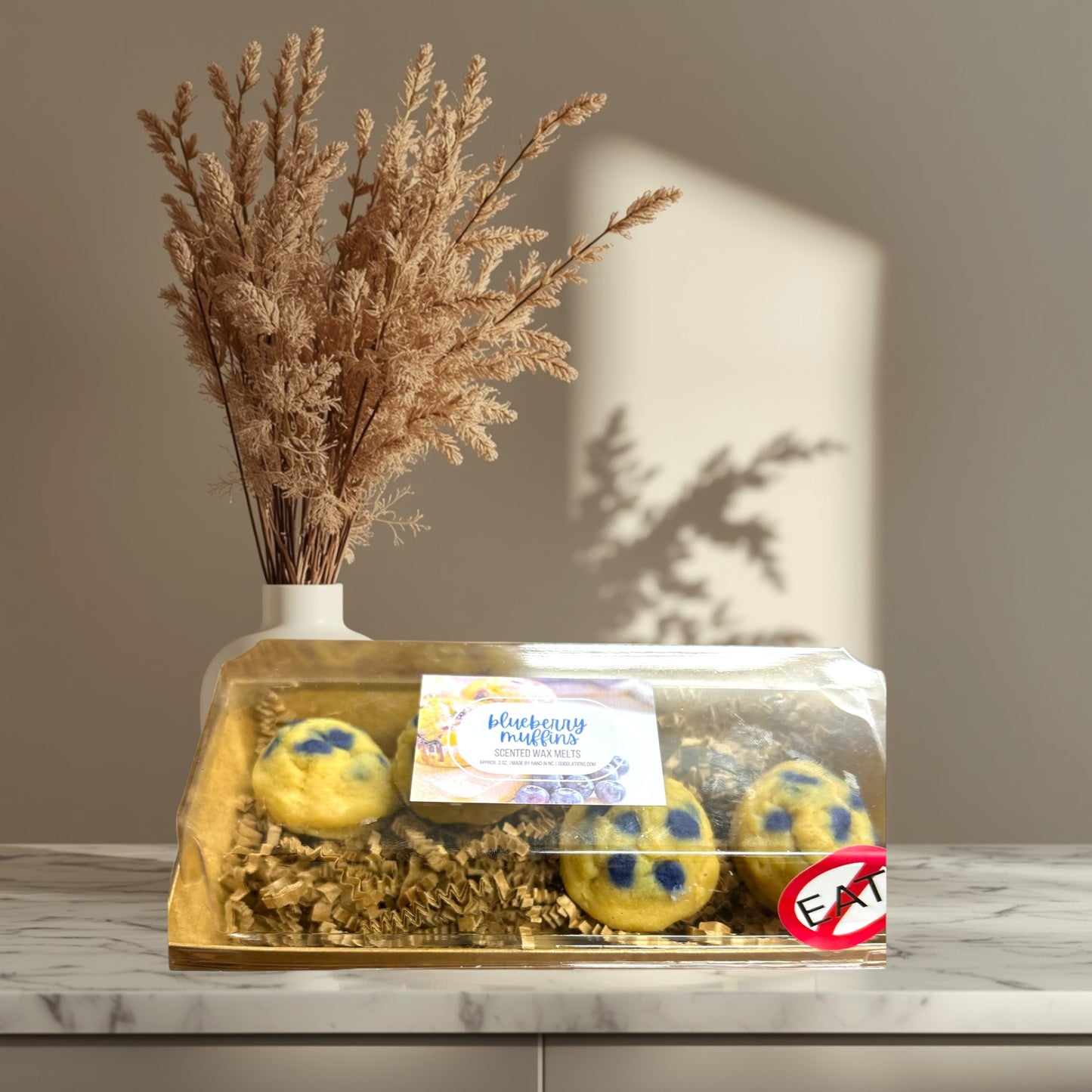 Doodlations Coffee Bar & Boutique Blueberry Muffin wax melts in a clear box with shredded paper, set on a striped cloth.