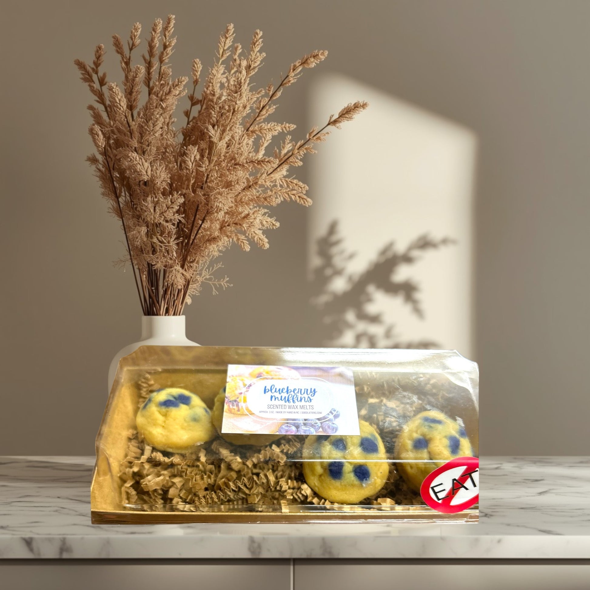 Doodlations Coffee Bar & Boutique Blueberry Muffin wax melts in a clear box with shredded paper, set on a striped cloth.