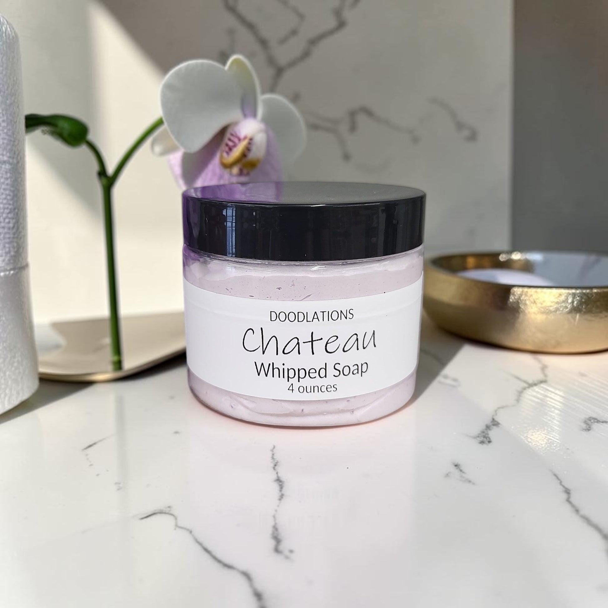 Doodlations Coffee Bar & Boutique Chateau whipped soap in a 4oz jar with a purple bath loofah on a marble surface.