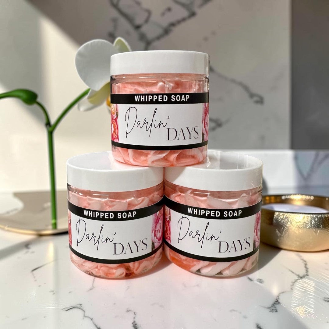 Doodlations Coffee Bar & Boutique Darlin Days whipped soap in clear jars with pink floral labels against greenery.