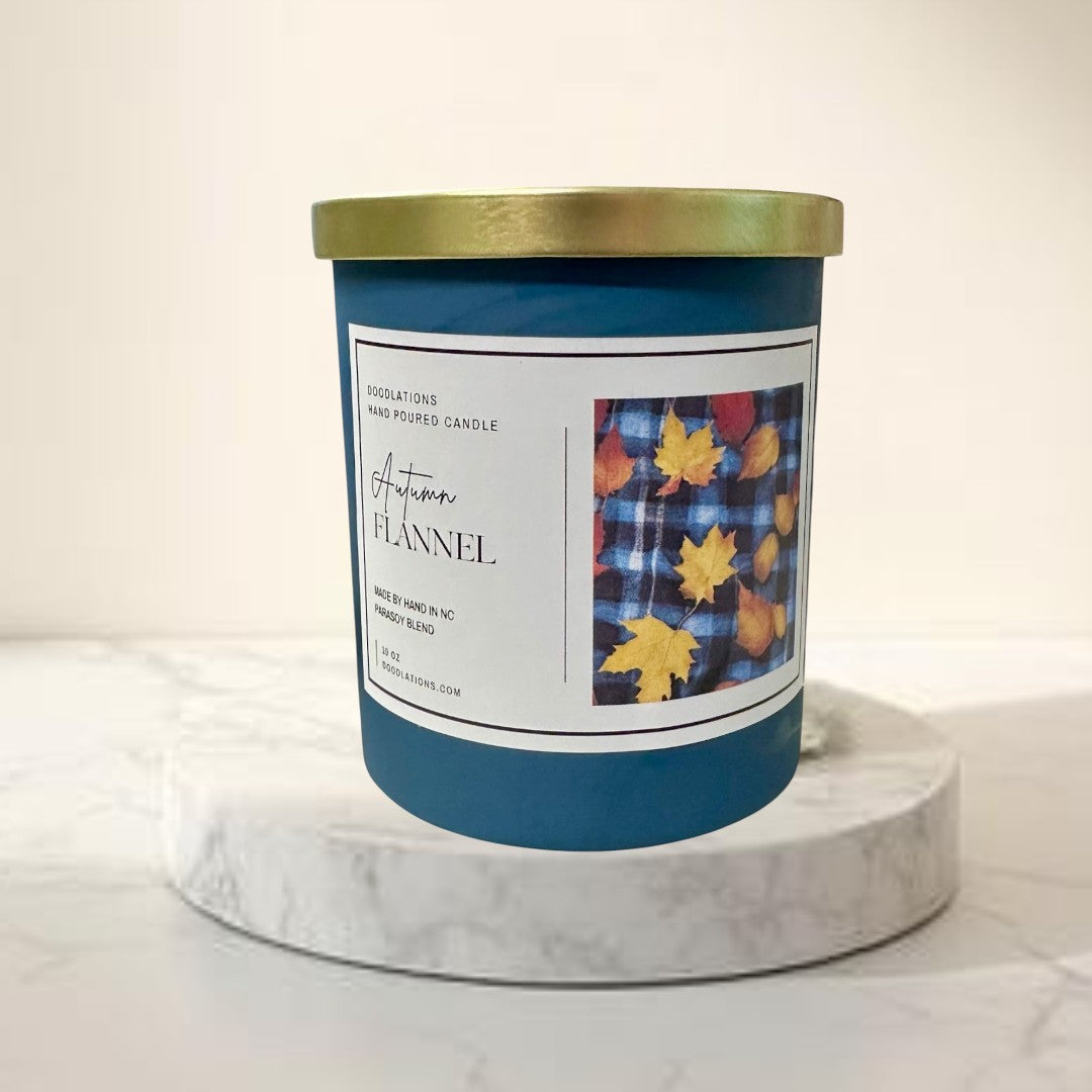 Doodlations Autumn Flannel luxury hand-poured soy blend candle in a teal jar with a gold lid. Features a label with plaid and fall leaves.