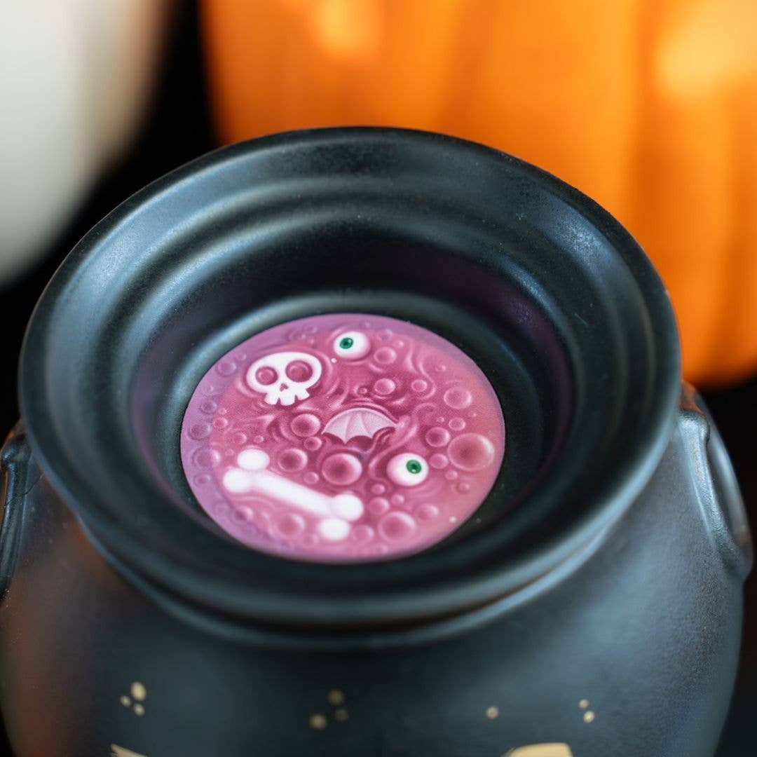 Doodlations Coffee Bar & Boutique matte black cauldron wax warmer with a melting pink wax disc featuring spooky icons.