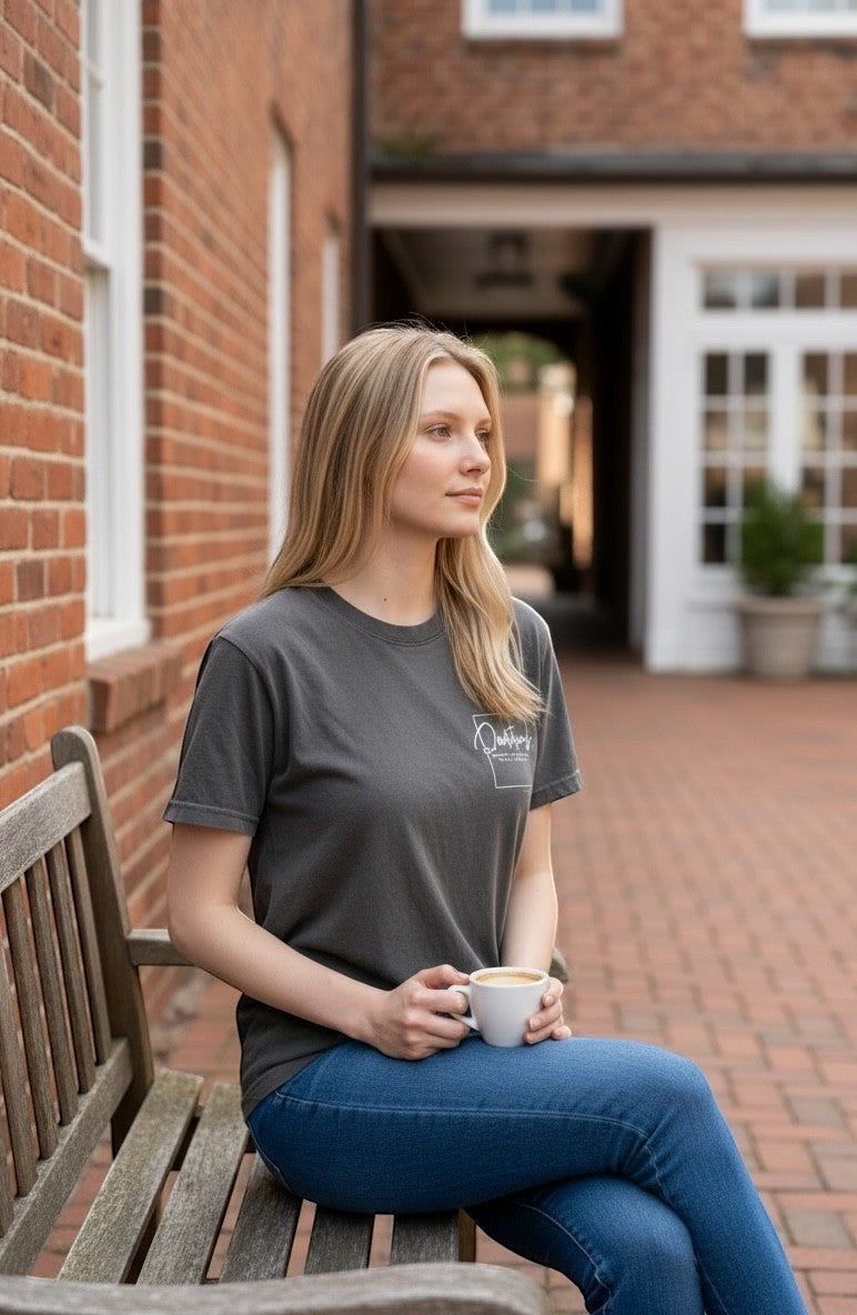 Woman wearing a charcoal grey Doodlations Coffee Bar & Boutique graphic tee while sitting on a bench with coffee