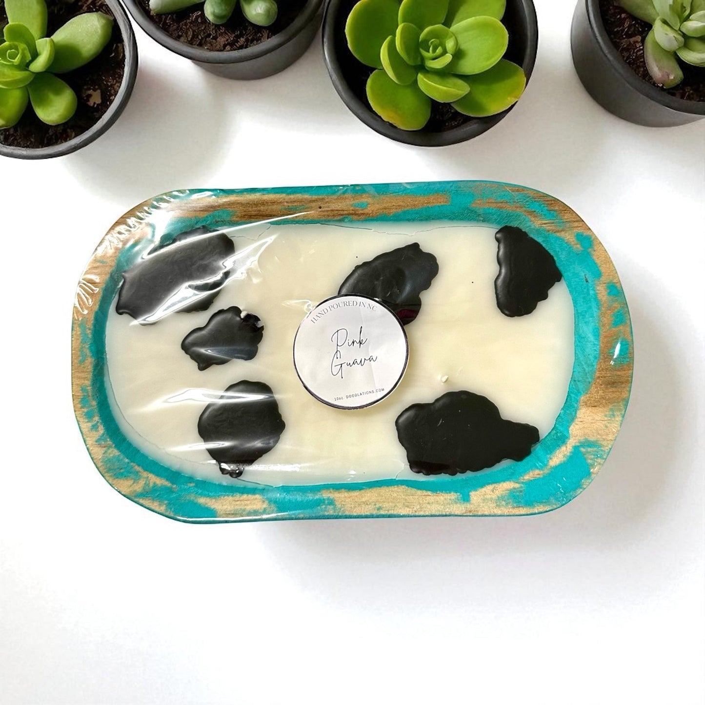 Doodlations Coffee Bar & Boutique cow dough bowl candle with pink guava scent, featuring black cow spots and a turquoise distressed wood finish.