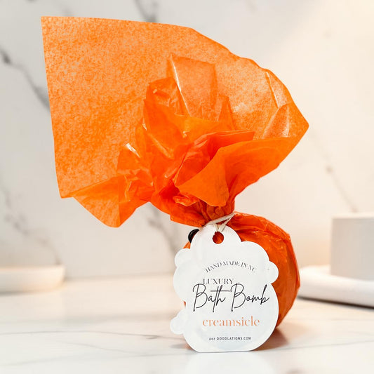Doodlations Coffee Bar & Boutique Creamsicle bath bomb wrapped in bright orange tissue paper with a product tag.