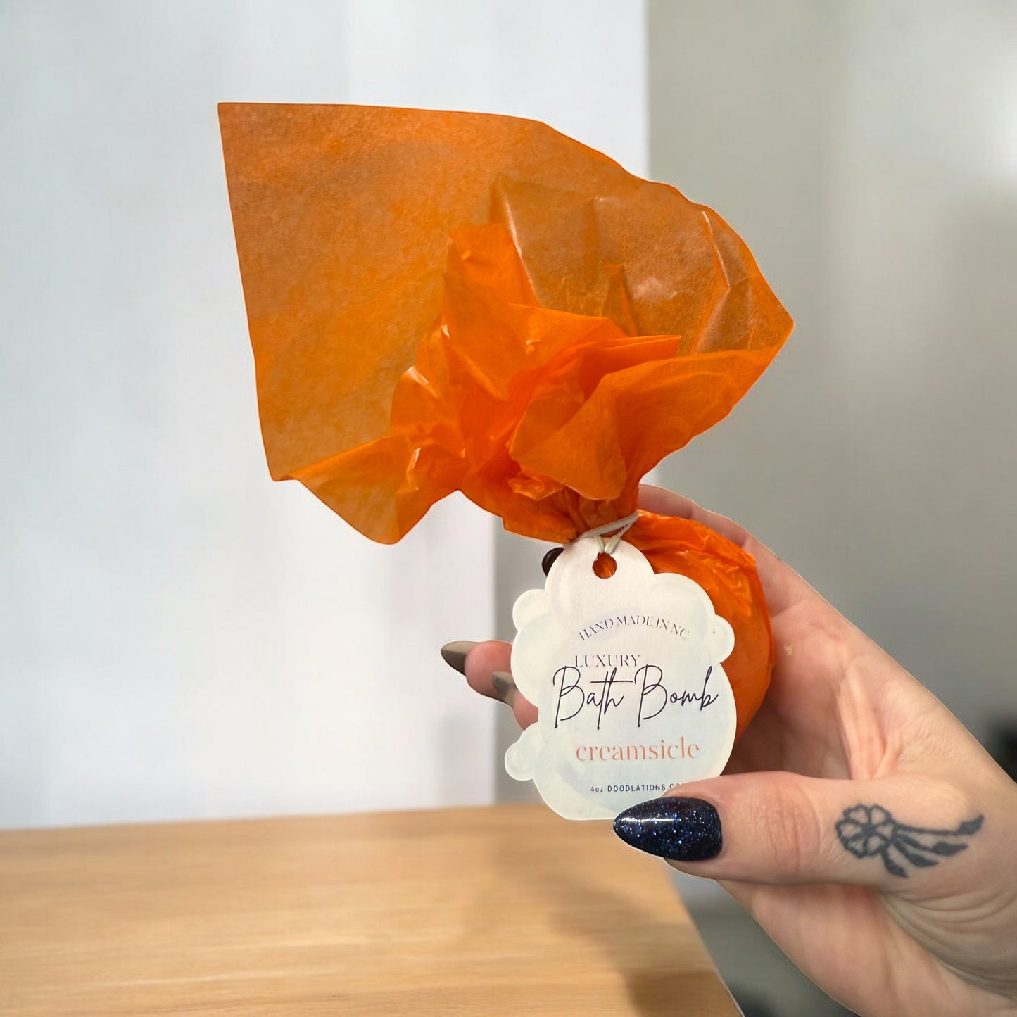 Doodlations Coffee Bar & Boutique Creamsicle bath bomb wrapped in orange tissue paper held in a hand