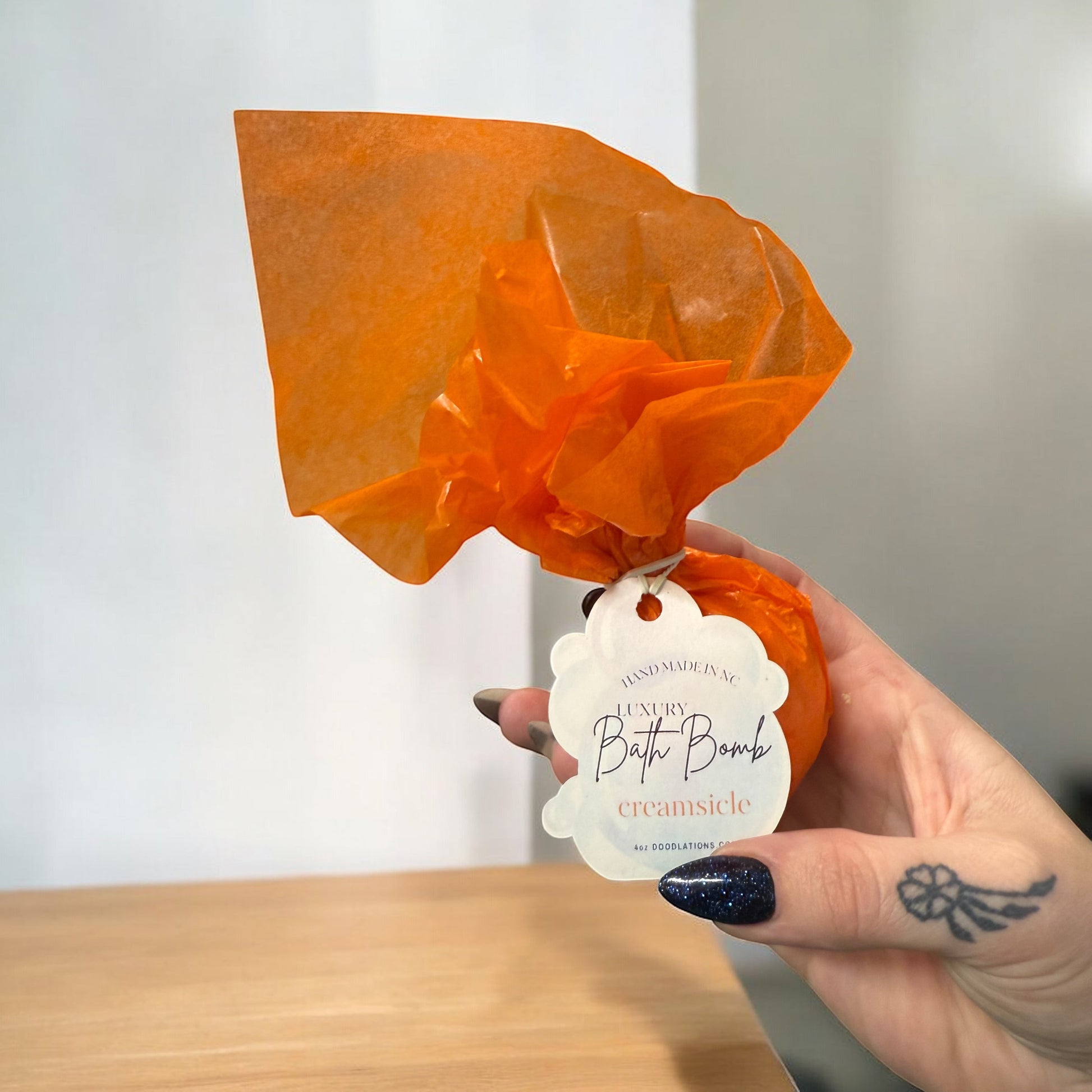Doodlations Coffee Bar & Boutique Creamsicle bath bomb wrapped in orange tissue paper held in a hand