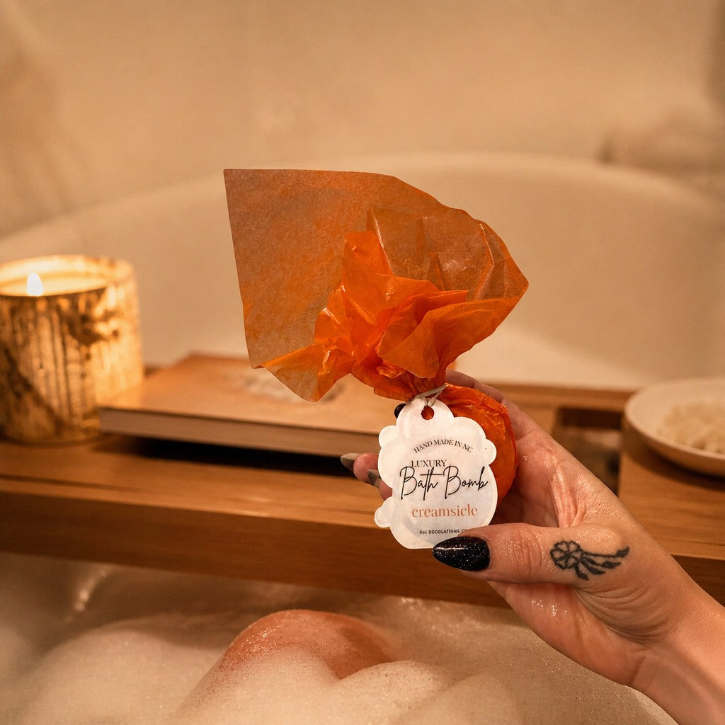 Doodlations Coffee Bar & Boutique Creamsicle bath bomb wrapped in orange paper held over a warm, relaxing bubble bath.
