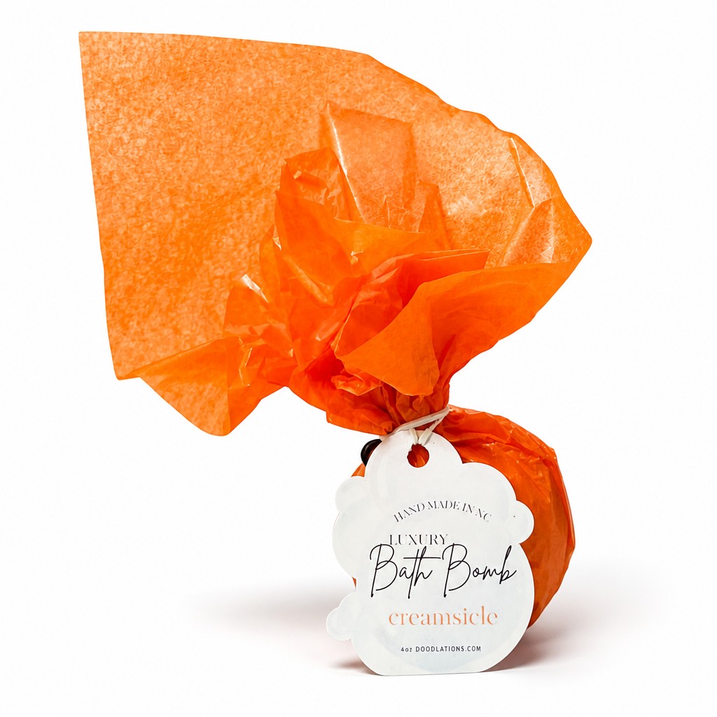 Doodlations Coffee Bar & Boutique Creamsicle bath bomb wrapped in bright orange tissue paper with a product tag.
