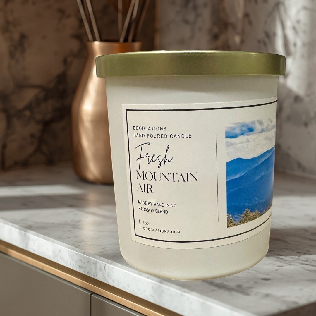 Doodlations Coffee Bar & Boutique Fresh Mountain Air hand-poured soy blend candle in a white jar with a gold lid.