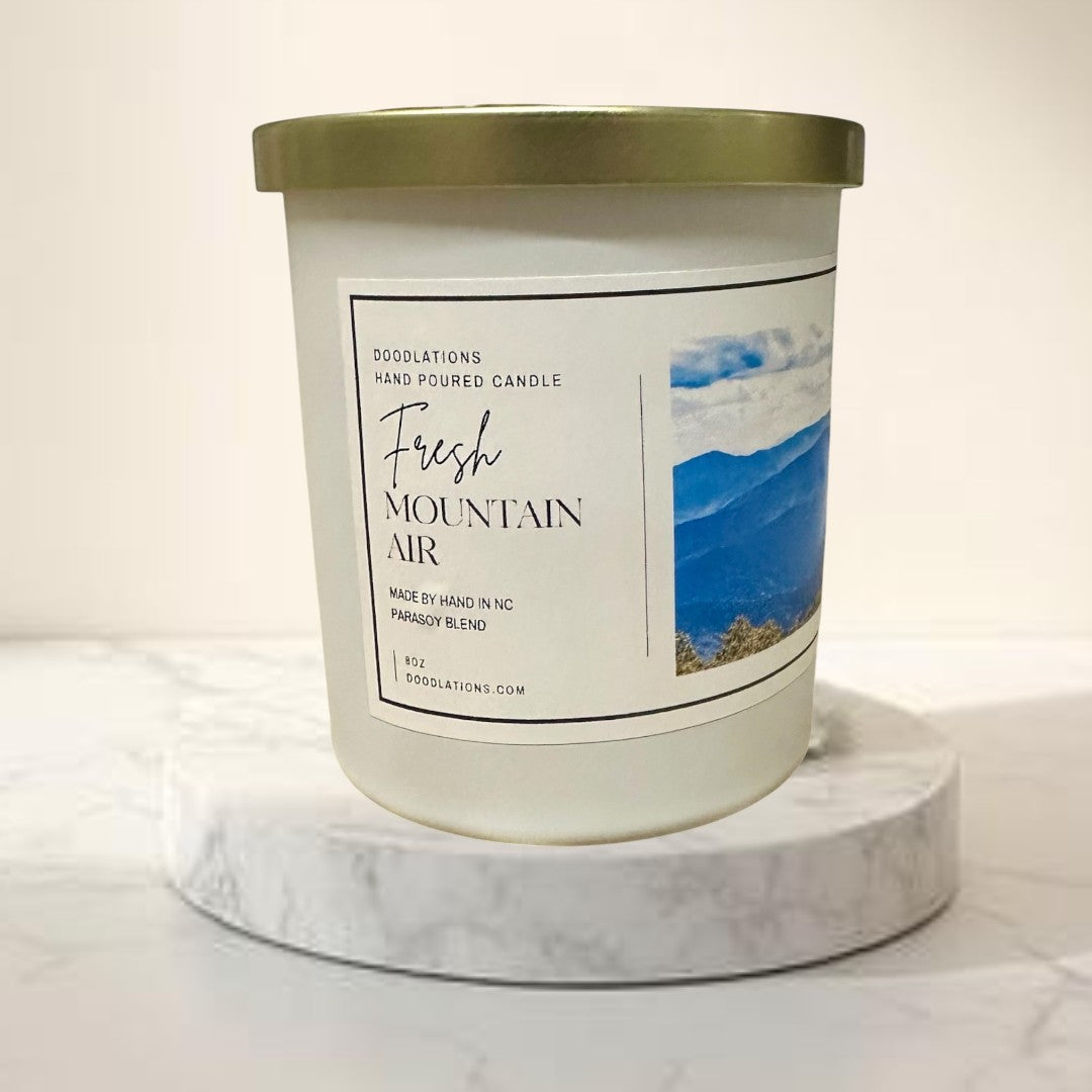 Doodlations Coffee Bar & Boutique Fresh Mountain Air hand-poured soy blend candle in a white jar with a gold lid.