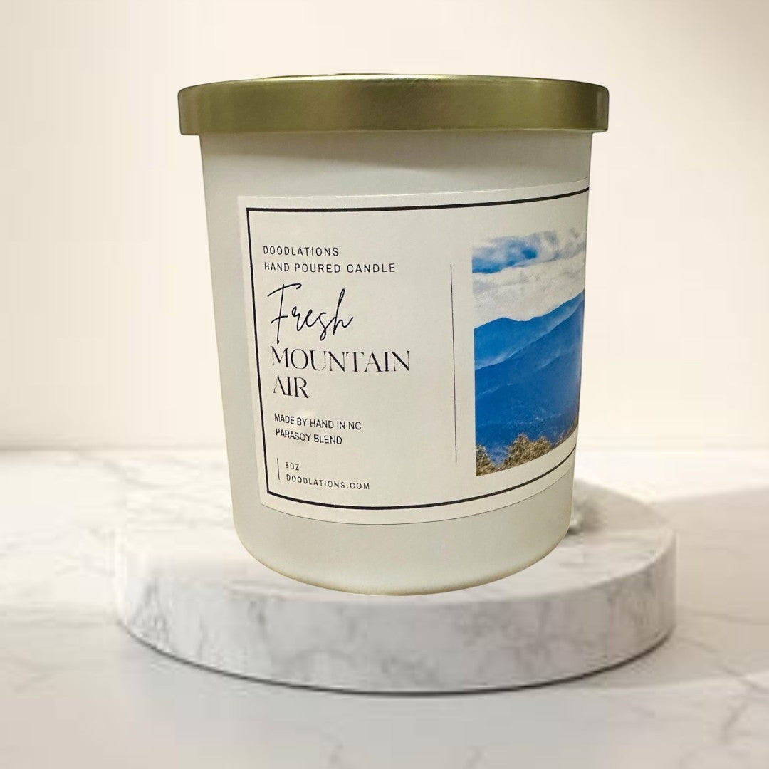 Doodlations Coffee Bar & Boutique Fresh Mountain Air hand-poured soy blend candle in a white jar with a gold lid.