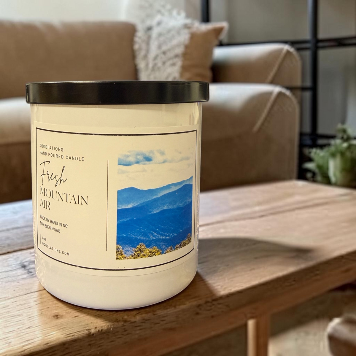 Doodlations Coffee Bar & Boutique Fresh Mountain Air soy blend candle in a white jar with a black lid on a wooden table